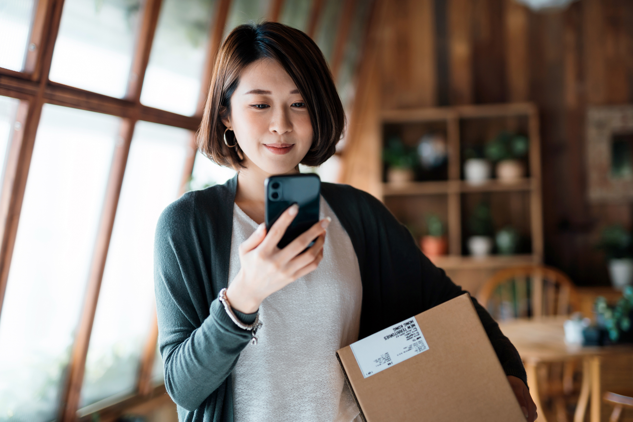 A woman smiling at her phone with a delivered parcel under her arm, standing by a sunny window at home.