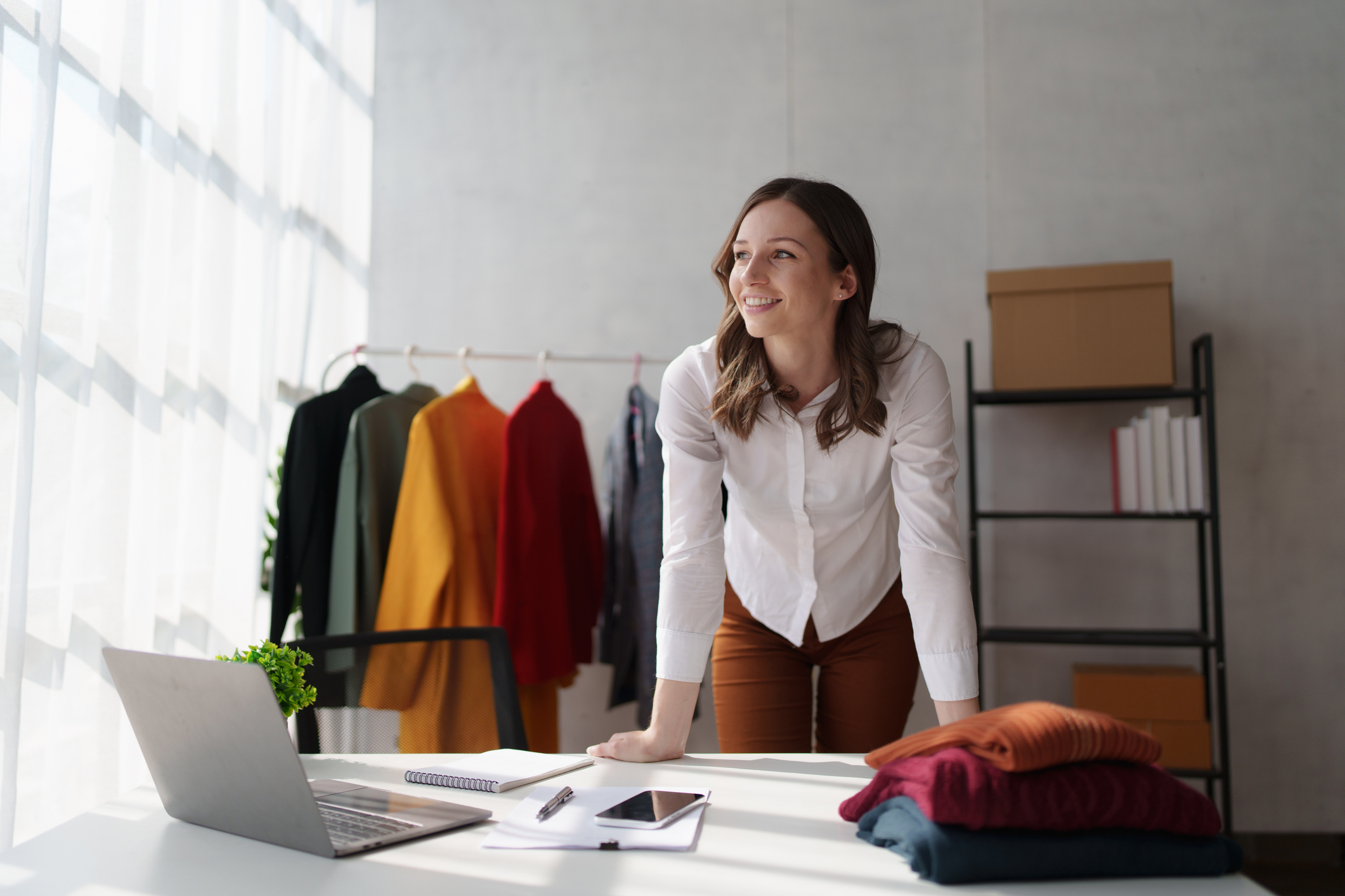 A small clothing-brand owner leaning on her worktable in her studio, smiling toward the window