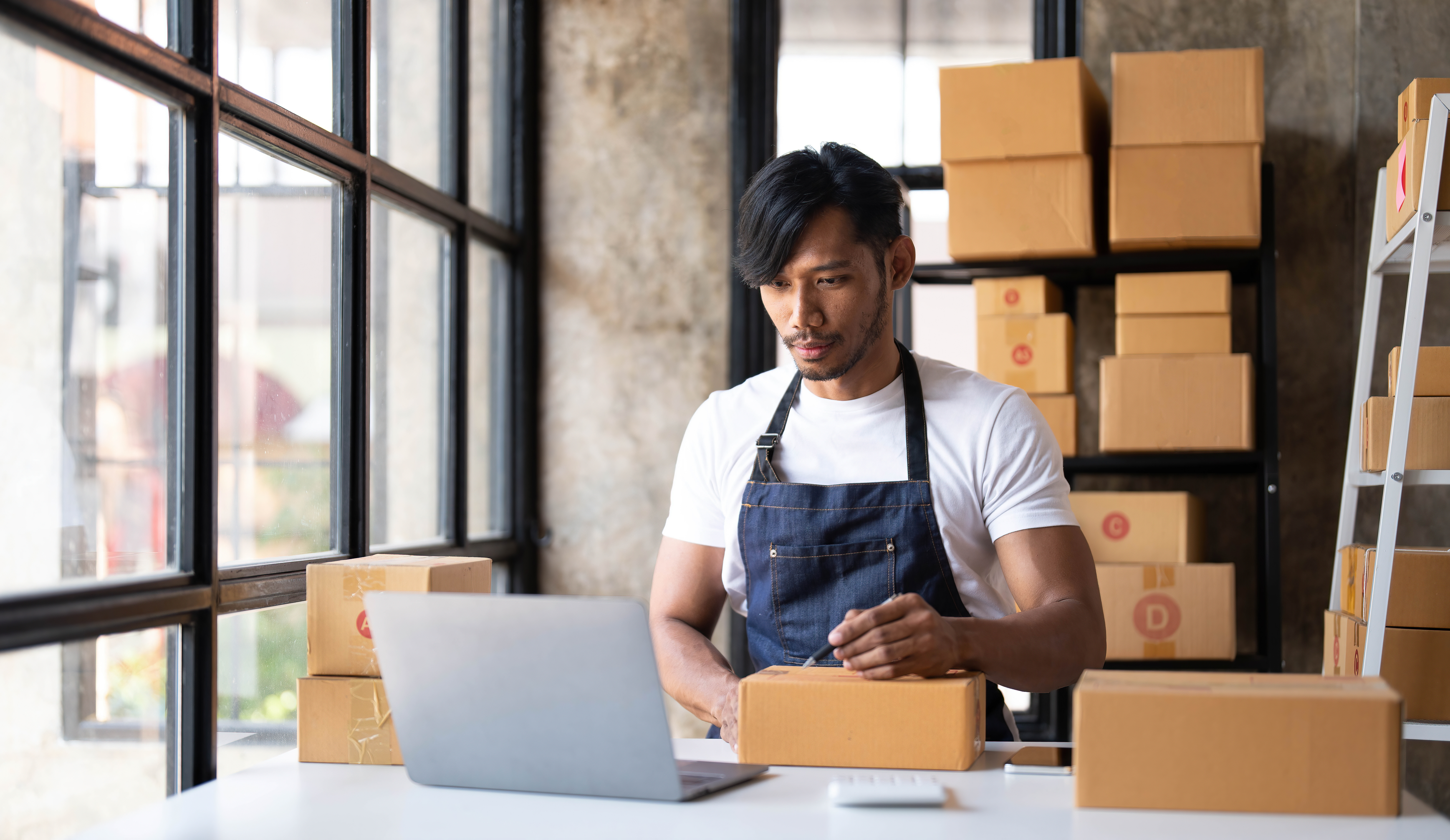 Small business operator preparing a shipment, writing an address on a box while reviewing online orders.