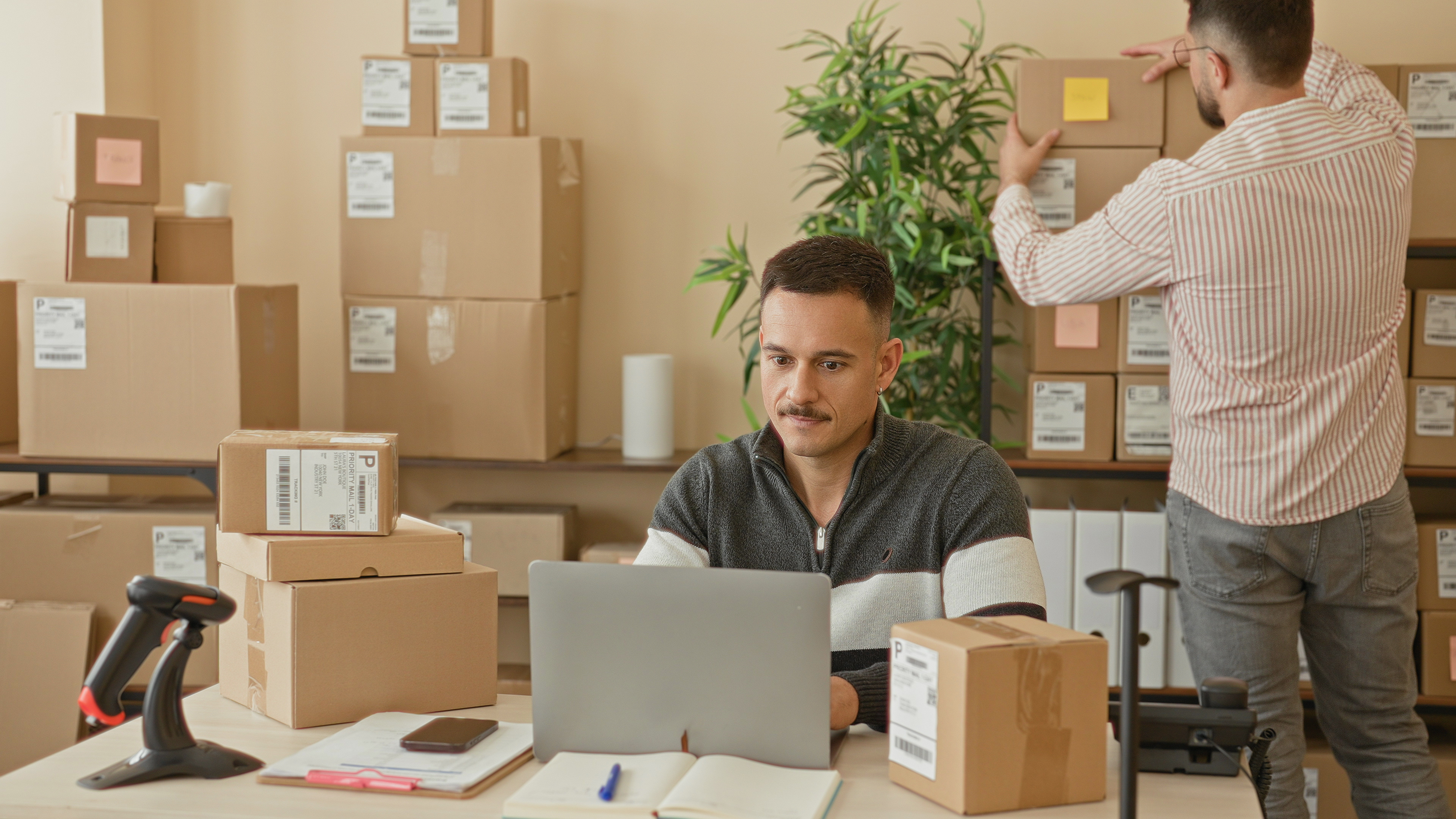 Two workers pointing at a laptop while packing boxes in a small business shipping area.